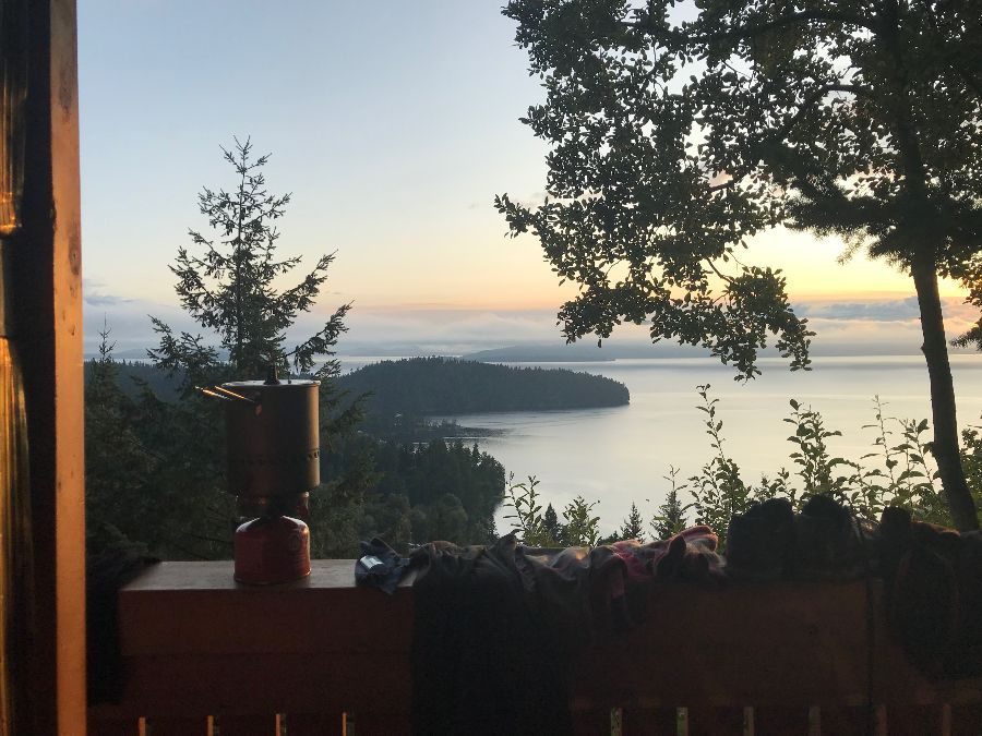 A camp stove and drying clothes on a cabin deck railing, above a bay at dusk.