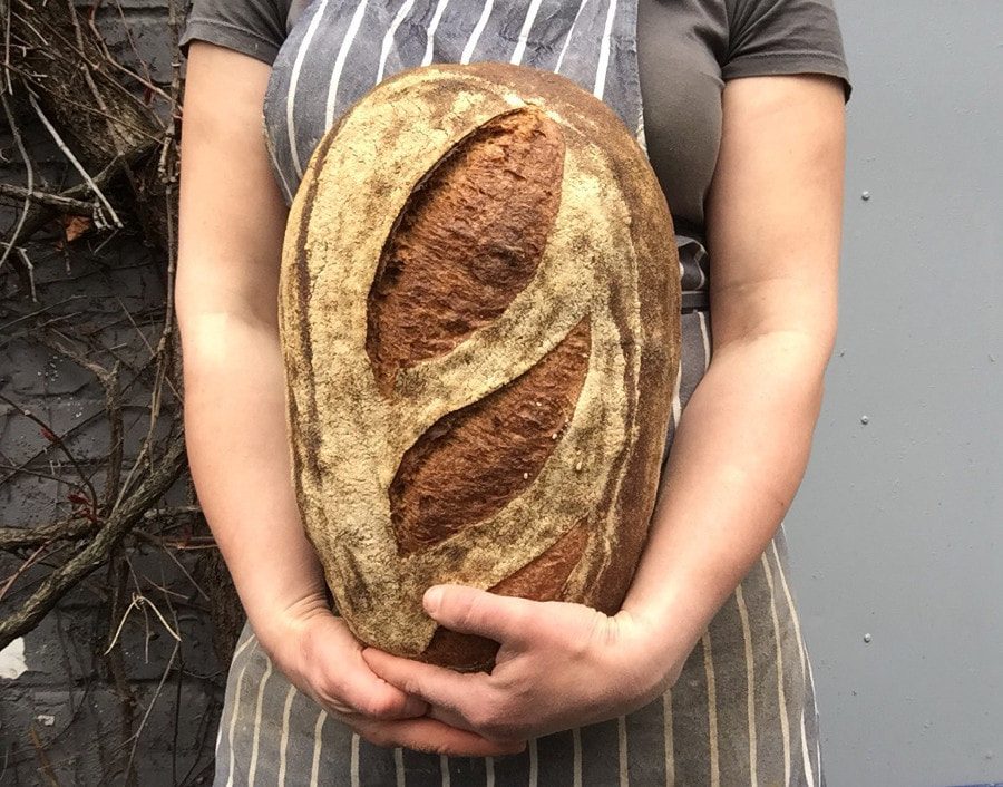 A baker in a striped apron holds a large, deeply scored loaf.